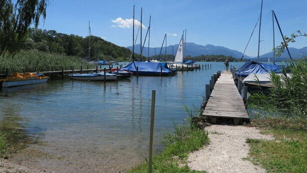 A calm lake with boats docked at a wooden pier, surrounded by greenery and mountains in the background.