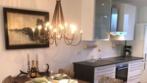 A dining area with a wooden table, chairs, and a chandelier. There is a kitchen in the background with white cabinets and a coffee maker.