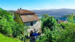 A two-story house with a stone base and a yellow upper section, surrounded by lush greenery and mountains in the background. - Villa Garibaldino (Podenzana)