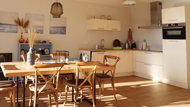 A modern kitchen with wooden cabinets, a dining table with chairs, and a hanging light fixture. - Sunny Apartment Directly on the Heegermeer (Heeg)