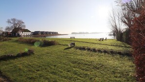 A grassy area with a bench and a cow, a body of water, and a row of houses in the background. - Sunny Apartment Directly on the Heegermeer (Heeg)
