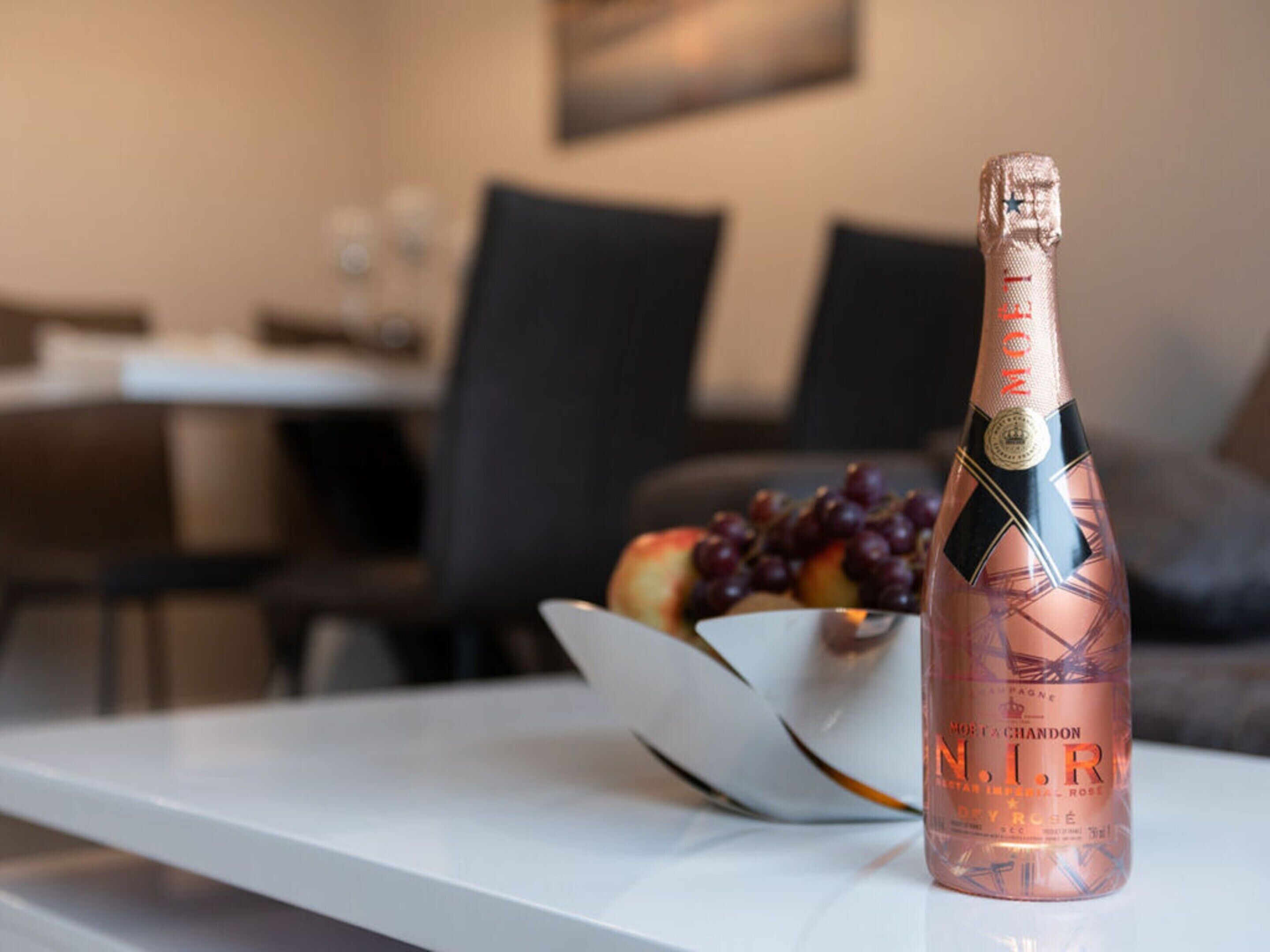 Une bouteille de champagne N.I.R. est posée sur une table blanche, avec un saladier de fruits et des grappes de raisin en arrière-plan.