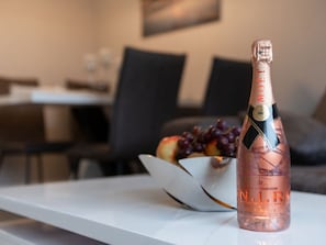 Une bouteille de champagne N.I.R. est posée sur une table blanche, avec un saladier de fruits et des grappes de raisin en arrière-plan.