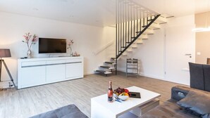 A modern living room with a white entertainment unit, a flat-screen TV, a staircase, and a seating area with a sofa and a coffee table. - Maiwaldo House Comfortable Vacation Home (Wurster Nordseeküste)