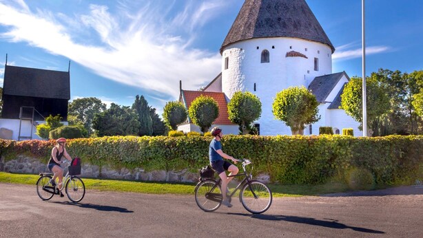 Two cyclists on a paved path with a historic tower and white building in the background. - 5 Person Holiday Park Home in Gudhjem (Gudhjem)