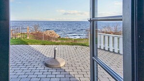 A view from a window overlooking a paved area with a stone structure, a white fence, and a rocky coastline. - 5 Person Holiday Park Home in Gudhjem (Gudhjem)