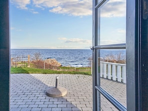 A view from a window overlooking a paved area with a stone structure, a white fence, and a rocky coastline. - 5 Person Holiday Park Home in Gudhjem (Gudhjem)