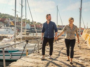 Un couple se promène sur une jetée en pierre, avec des bateaux et des bâtiments en arrière-plan.