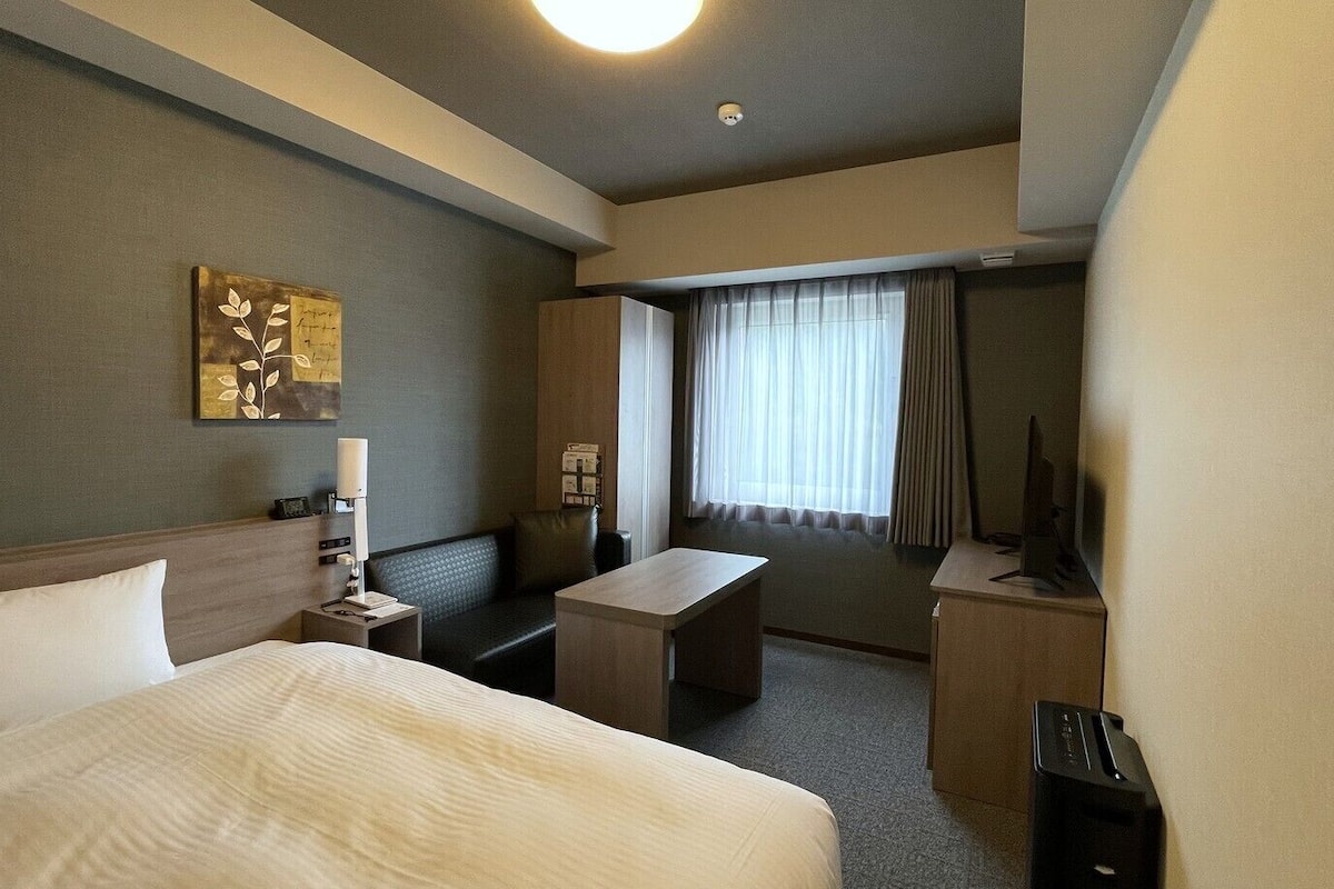 A hotel room with a bed, a desk, a chair, a TV, and a window with curtains.