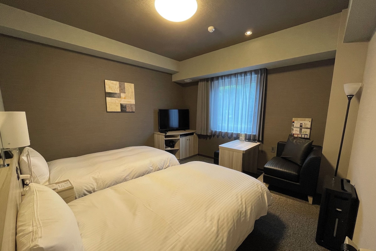 A hotel room with two beds, a desk, a television, a chair, and a lamp.