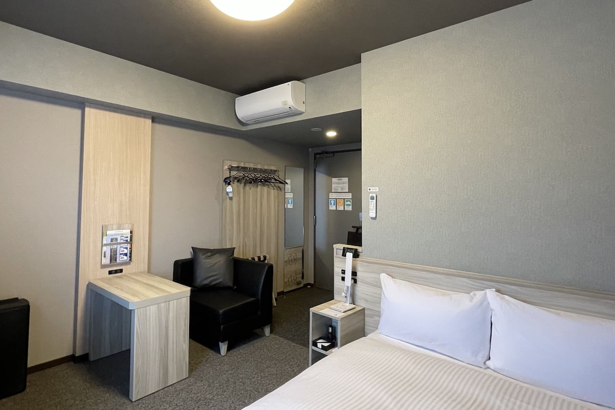A hotel room with a bed, a desk, a chair, a small table, an air conditioning unit, and a wall-mounted telephone.