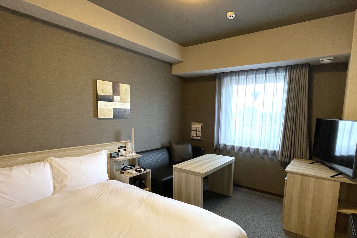 A hotel room with a large bed, a desk, a chair, a television, and a window with curtains.