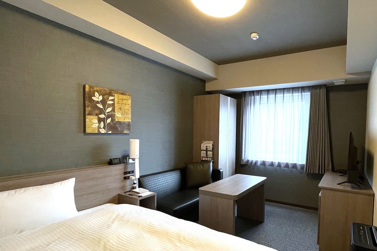 A hotel room with a bed, a desk, a chair, a small table, and a TV.