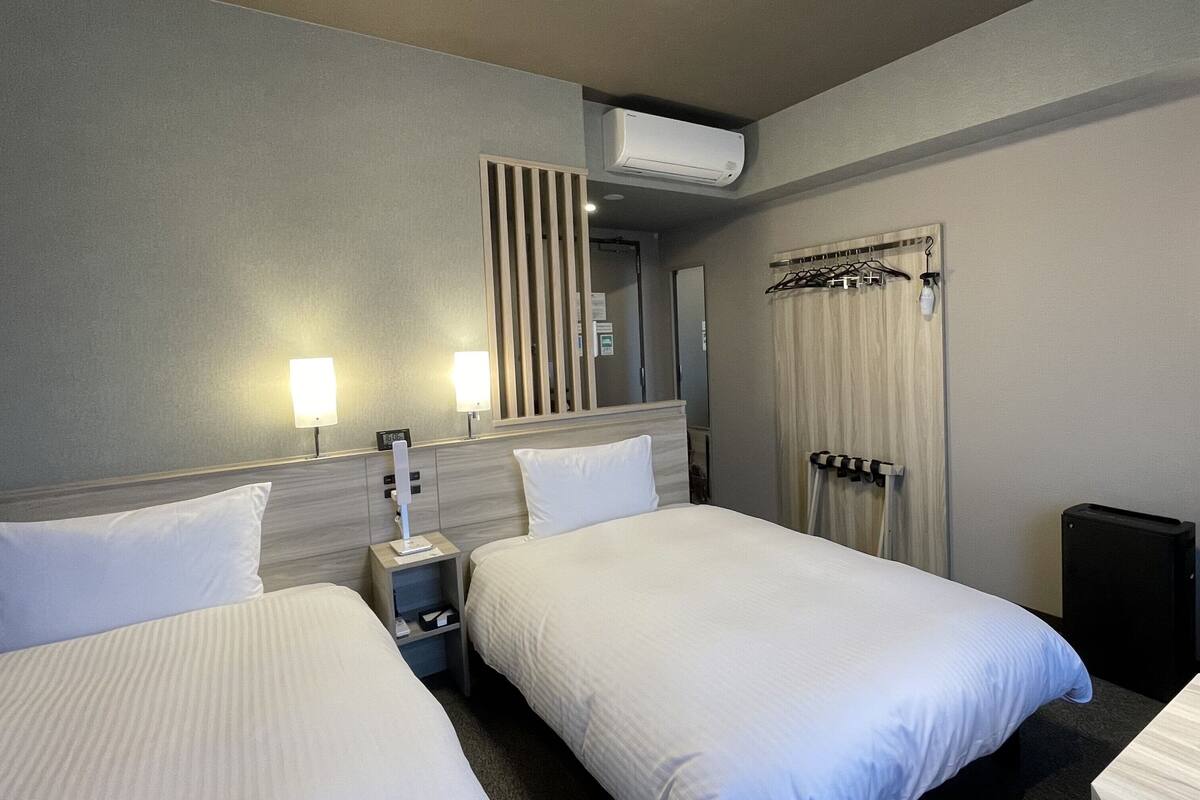 A hotel room with two beds, a bedside table with a lamp, a wall-mounted air conditioner, and a door with a towel rack.