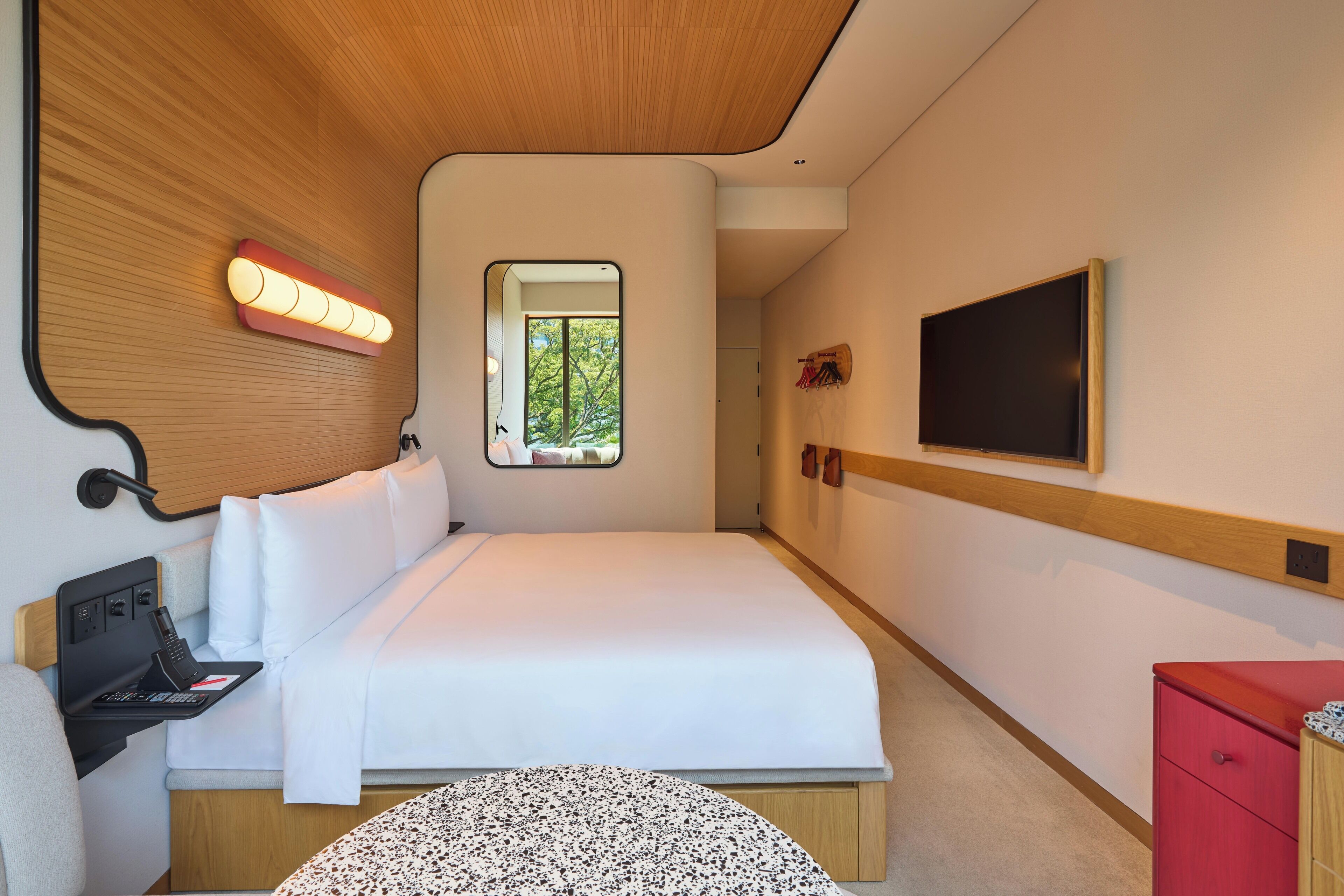 A modern hotel room with a bed, a small desk with a laptop, a TV mounted on the wall, and a window with a view of greenery.