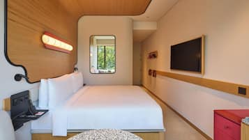 A modern hotel room with a bed, a small desk with a laptop, a TV mounted on the wall, and a window with a view of greenery.