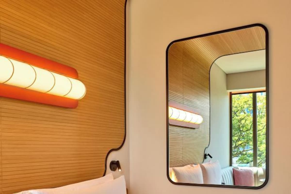 A hotel room with a wooden panel wall, a bedside lamp, a large mirror, and a window with a view of greenery.