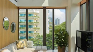 Ein modernes Wohnzimmer mit einer Couch, einem Couchtisch und einem Fernseher. Große Fenster bieten Blick auf Gebäude und Grünflächen.