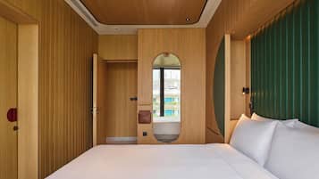 A hotel room with a wooden panel wall, a large bed with white linens, a small mirror, and a view of a building through a window.