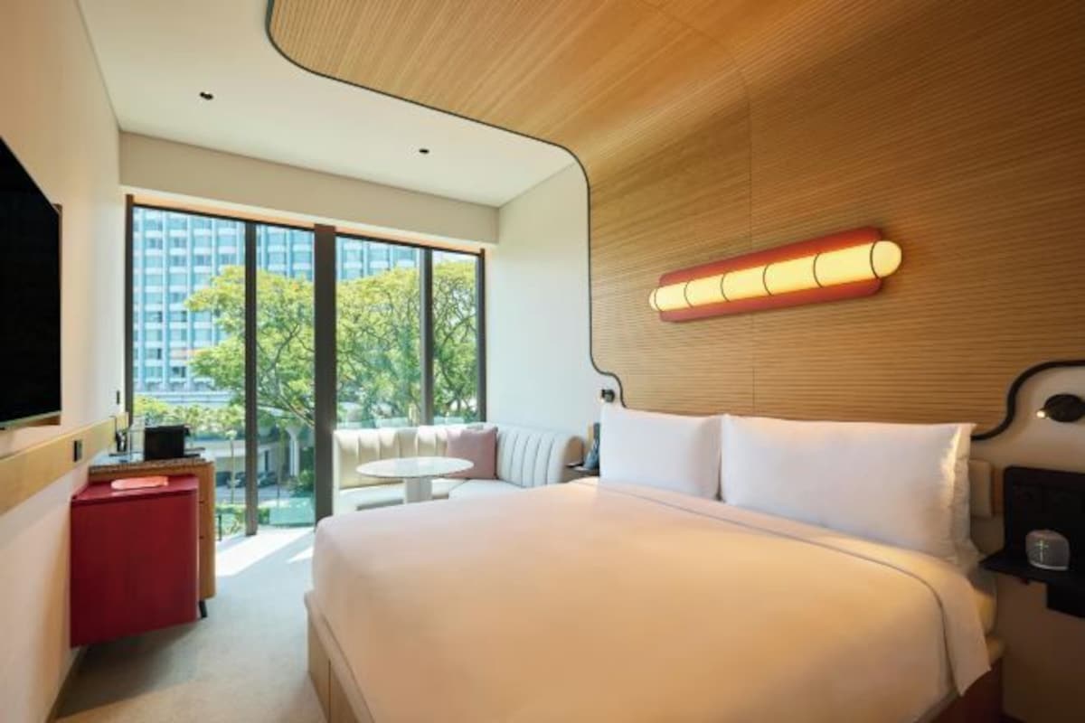 A modern hotel room with a large bed, a red side table, a round table, and a large window offering a view of buildings and trees.