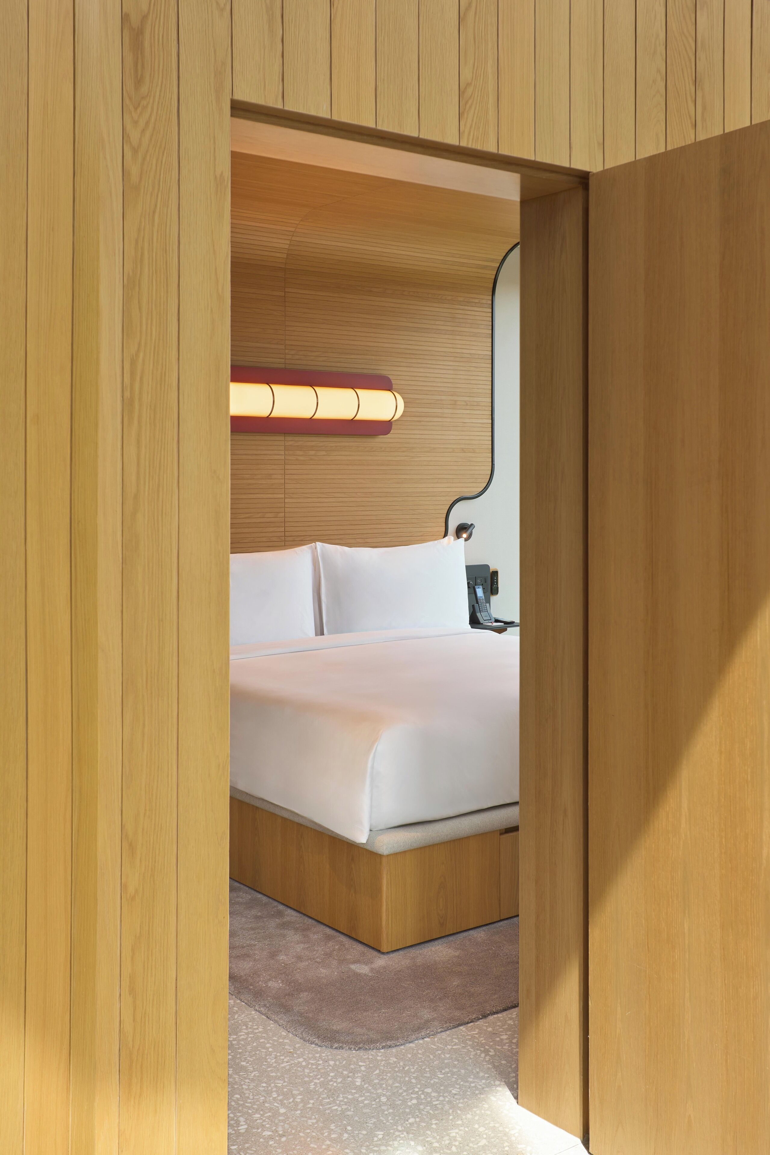 A hotel room with a wooden headboard, a bed with white linens, a bedside table with a lamp, and a carpeted floor.
