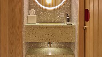 Un baño con tocador de madera, un lavamanos con encimera moteada, un espejo con luz y una caja de pañuelos.
