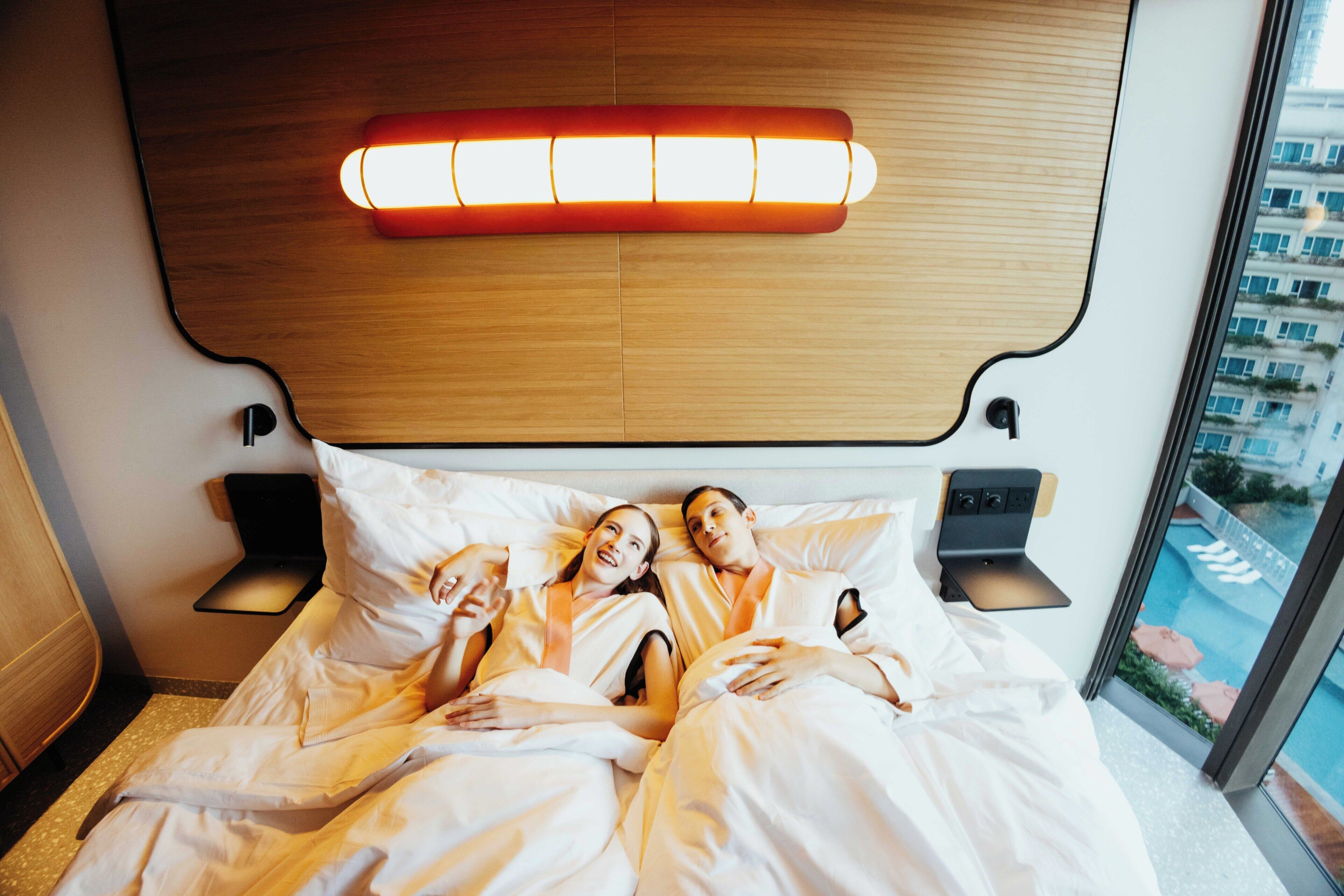 Une chambre d’hôtel avec un lit, des tables de chevet, une lampe fixée au mur et une vue sur les bâtiments par la fenêtre.
