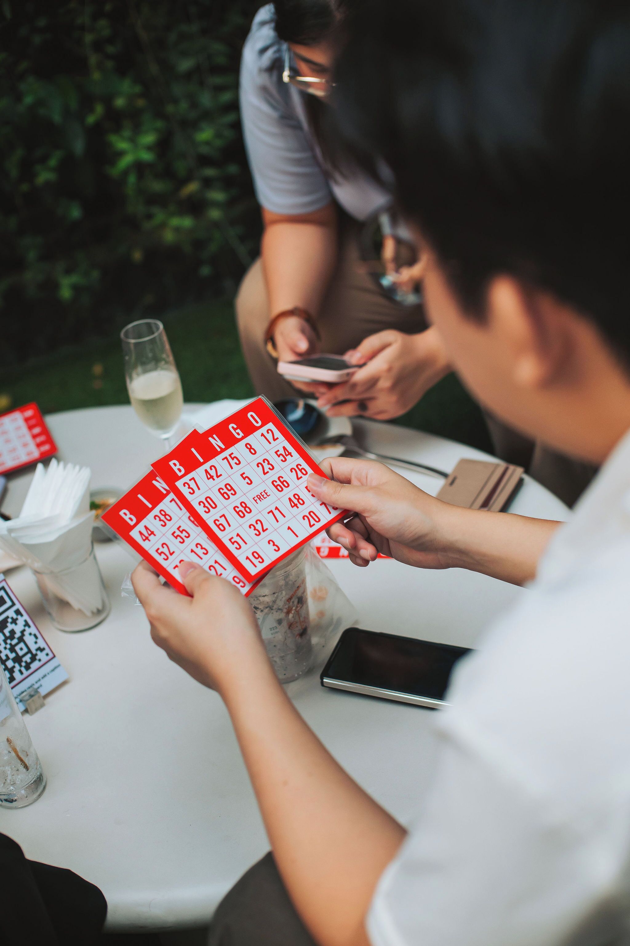 A person holding a bingo card with numbers visible, seated at a table with a smartphone, a glass of a clear liquid, and a QR code on a card.