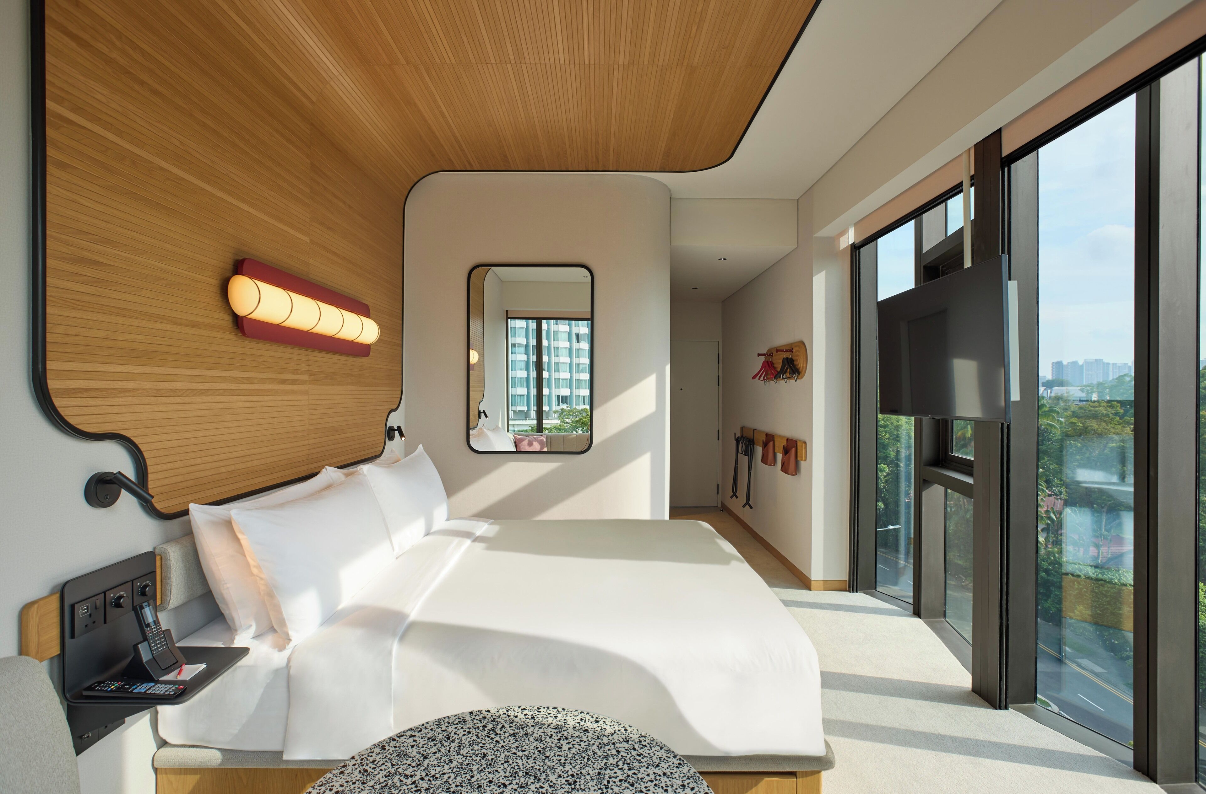 A modern bedroom with a large bed, a bedside table with a phone, a mirror, and a view of the city through large windows.