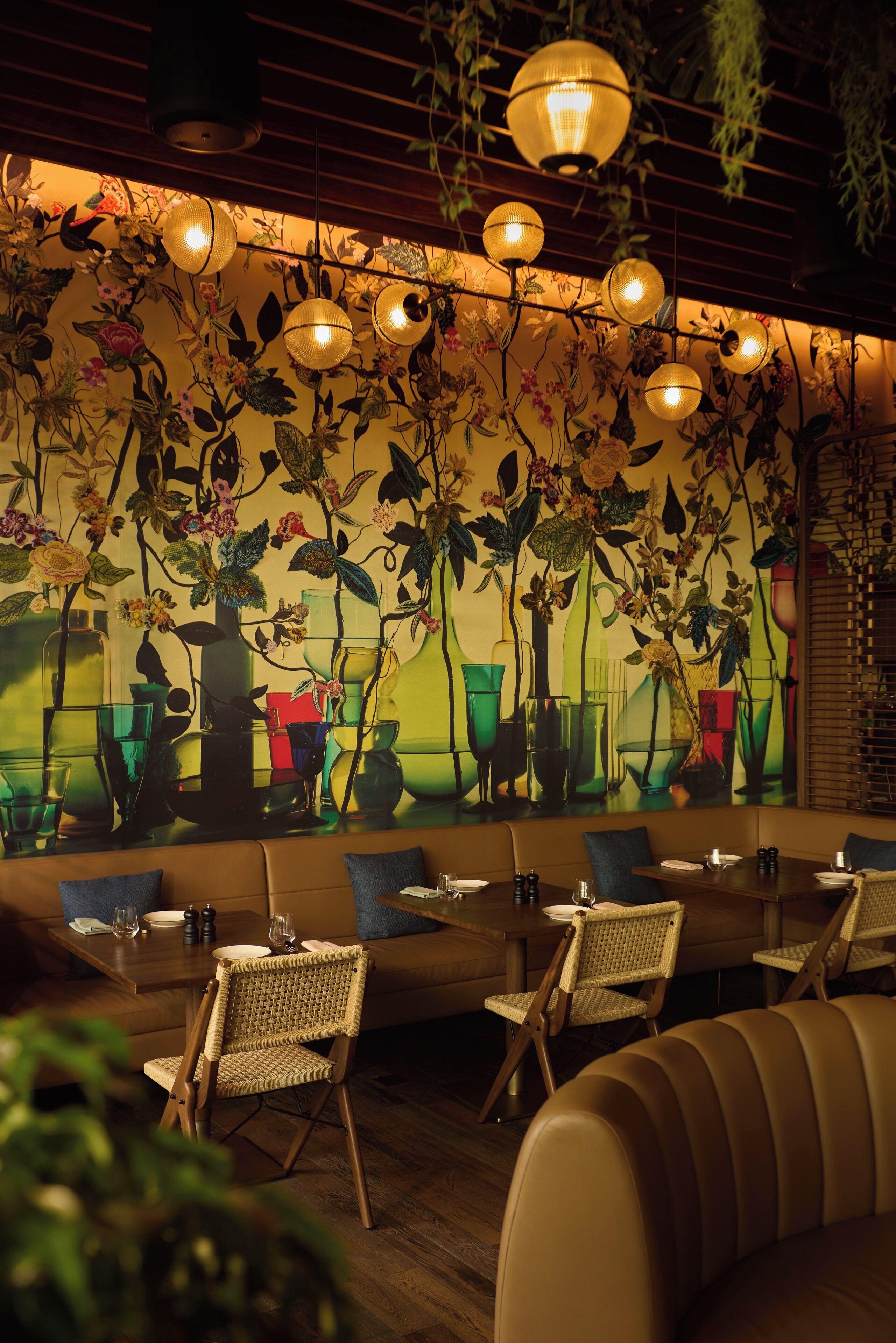 A restaurant interior with a mural of vases and flowers, wicker chairs, and a wooden table with plates and glasses.