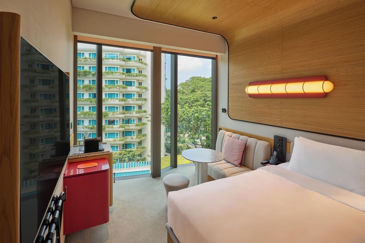 Une chambre d’hôtel moderne, dotée d’un grand lit, d’une petite table, d’une télévision et offrant une vue sur un immeuble avec des balcons.