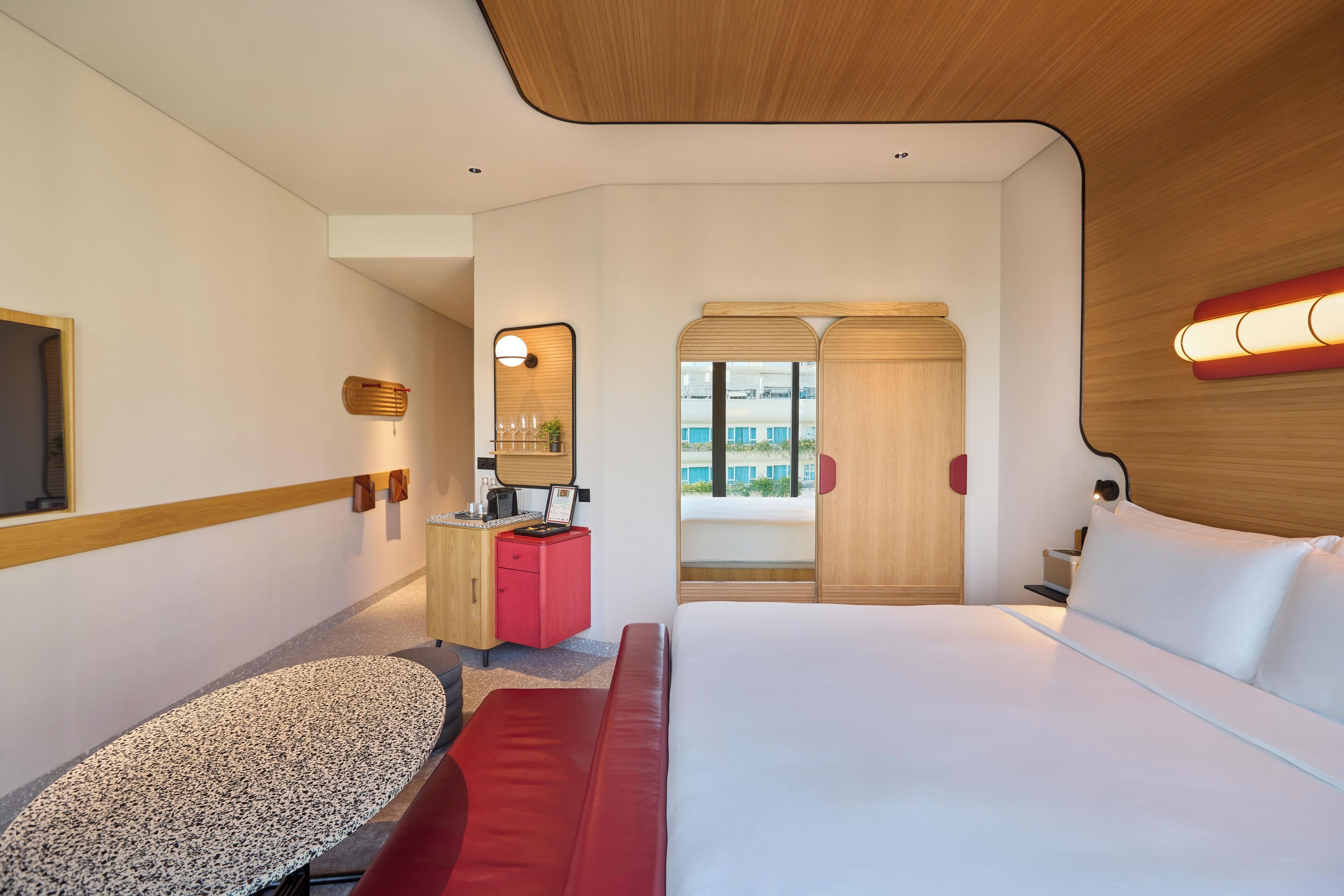 Una camera d'albergo con un letto, una scrivania, una sedia rossa, un tavolino e vista all'esterno.