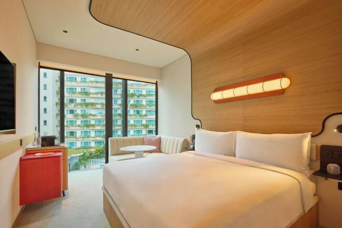 Une chambre d’hôtel moderne avec un grand lit, un petit bureau rouge, une table ronde et une vue sur un immeuble de grande hauteur.