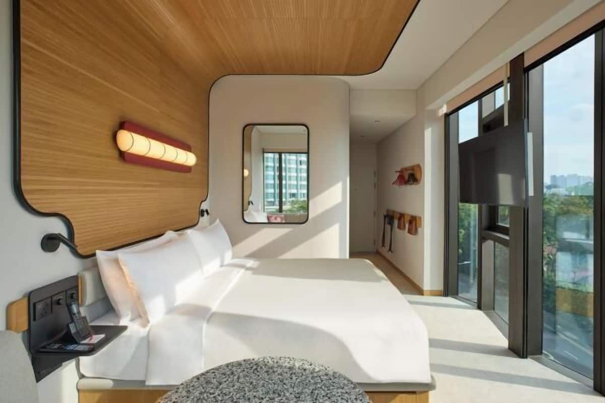Une chambre moderne avec un grand lit, une table de chevet avec un téléphone, un miroir et une vue sur la ville grâce à de grandes fenêtres.