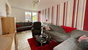 Ein modernes Wohnzimmer mit einem grauen Sofa, einem schwarzen Couchtisch und einem roten Teppich. Ein Fenster bietet Blick auf einen grünen Zaun und ein Gebäude.