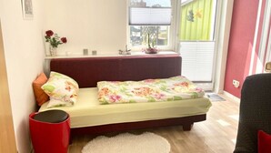 Ein kleines Schlafzimmer mit einem Bett, einer roten Mülltonne, einem weißen Teppich und einem Fenster mit Jalousien.
