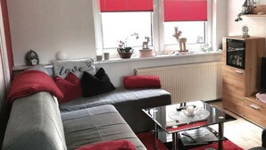 Ein modernes Wohnzimmer mit einem grauen Sofa, roten Kissen, einem Glastisch und einem Fenster mit rotem Rahmen.