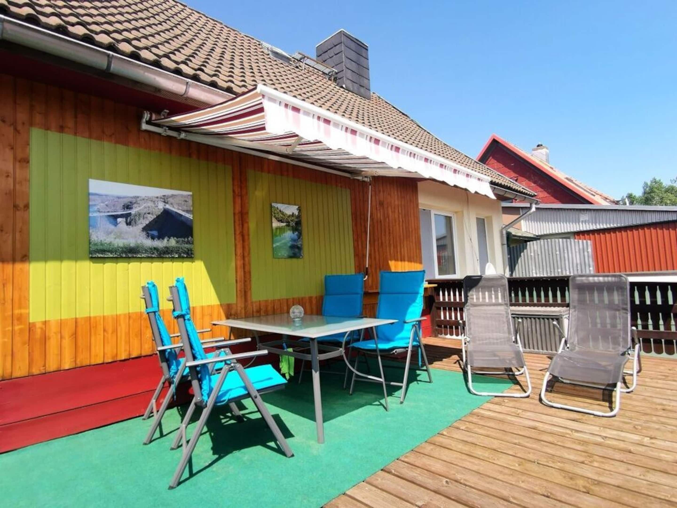 Un espacio al aire libre techado con mesa y sillas, una terraza de madera y un edificio de paredes coloridas y tejido de tejas.