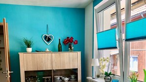 Ein Zimmer mit türkisfarbenen Wänden, einem Holzschrank mit Glastüren, einer herzförmigen Dekoration, einer Flasche, roten Blumen und einem Fenster mit Jalousien.