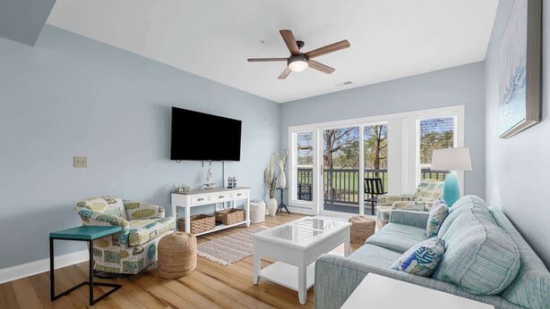 A living room with a ceiling fan, a flat-screen TV, a sofa, a coffee table, and a window with a view of the outdoors. - Golf Links Residence 3BR (Myrtle Beach)