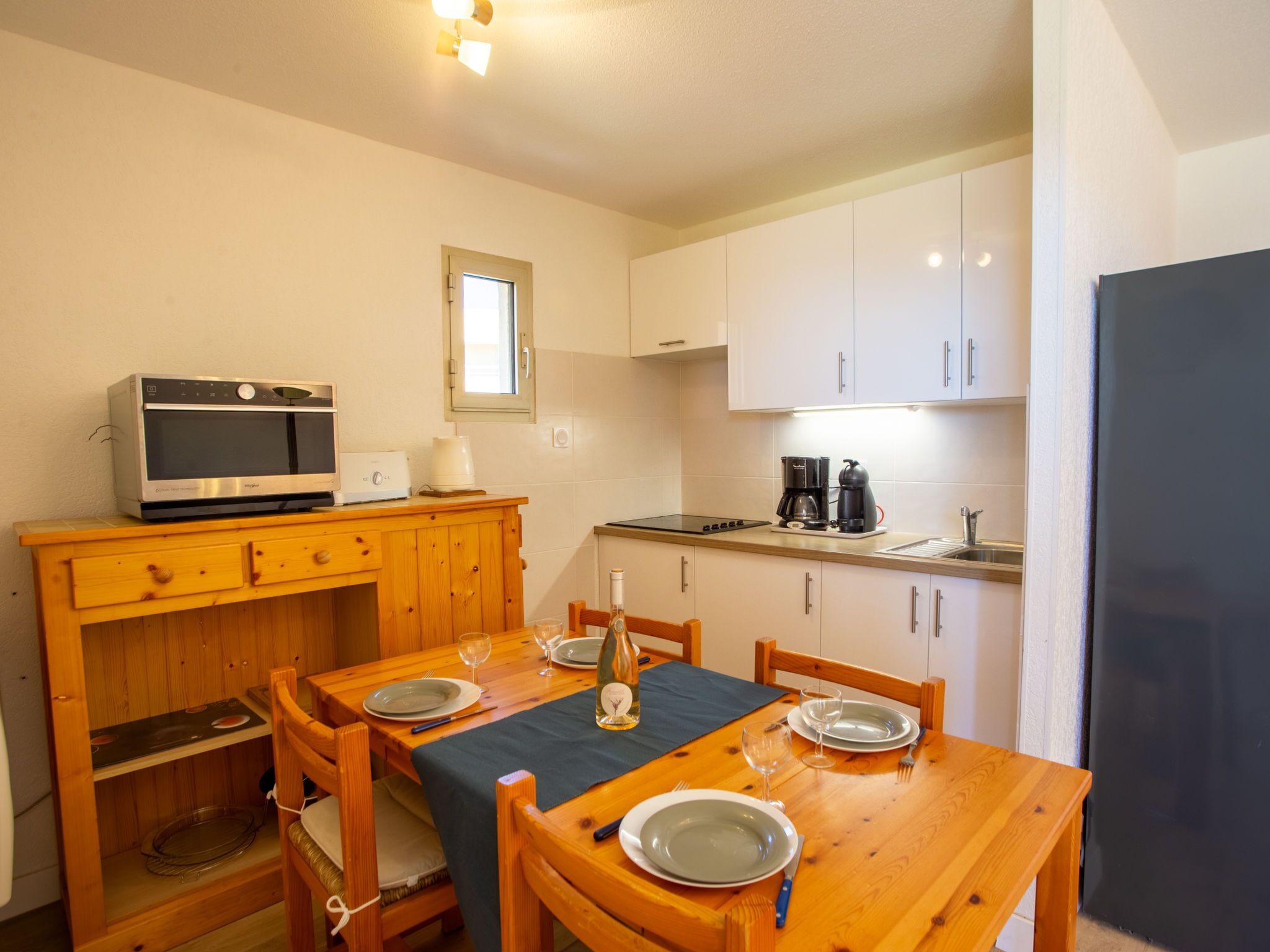 Una cocina compacta con armarios blancos, una mesa de comedor de madera preparada para dos comensales y un horno empotrado.