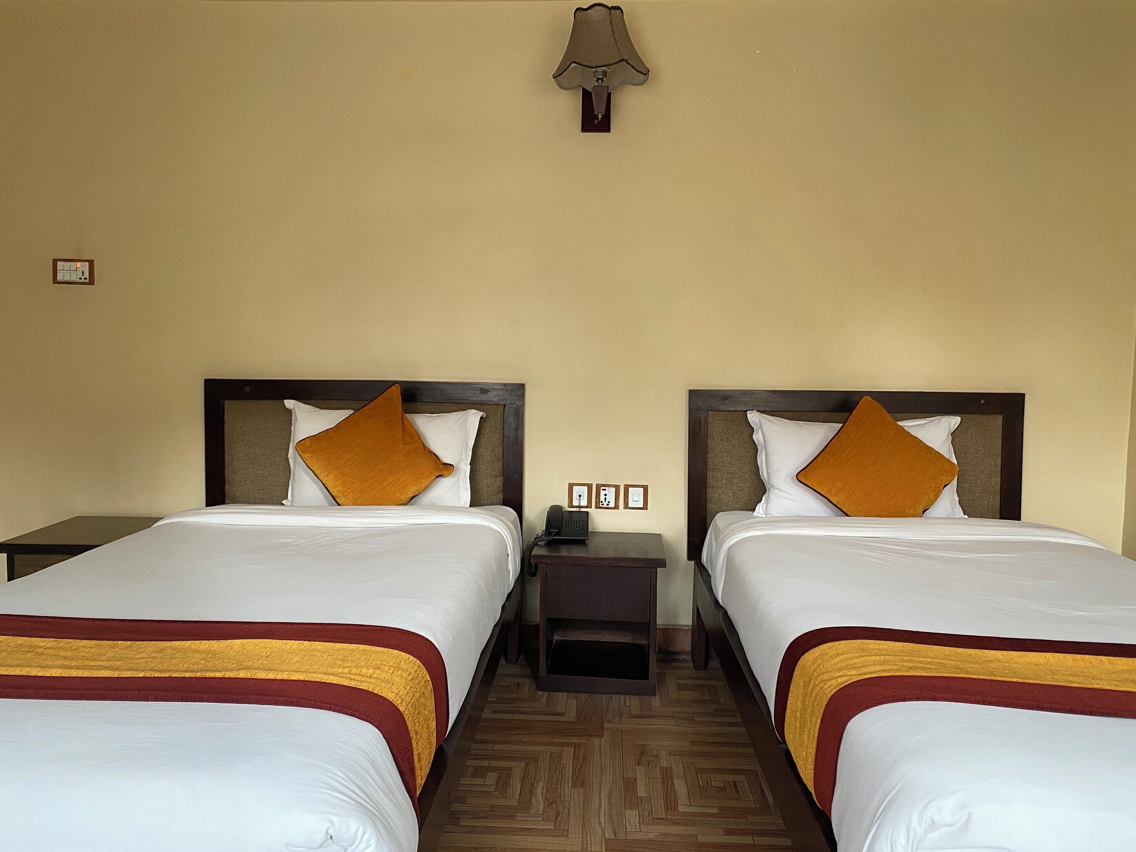 A hotel room with two beds, each with a wooden headboard and a brown accent pillow. There is a nightstand between the beds with a lamp and a phone. The room has a wooden parquet floor and beige walls.