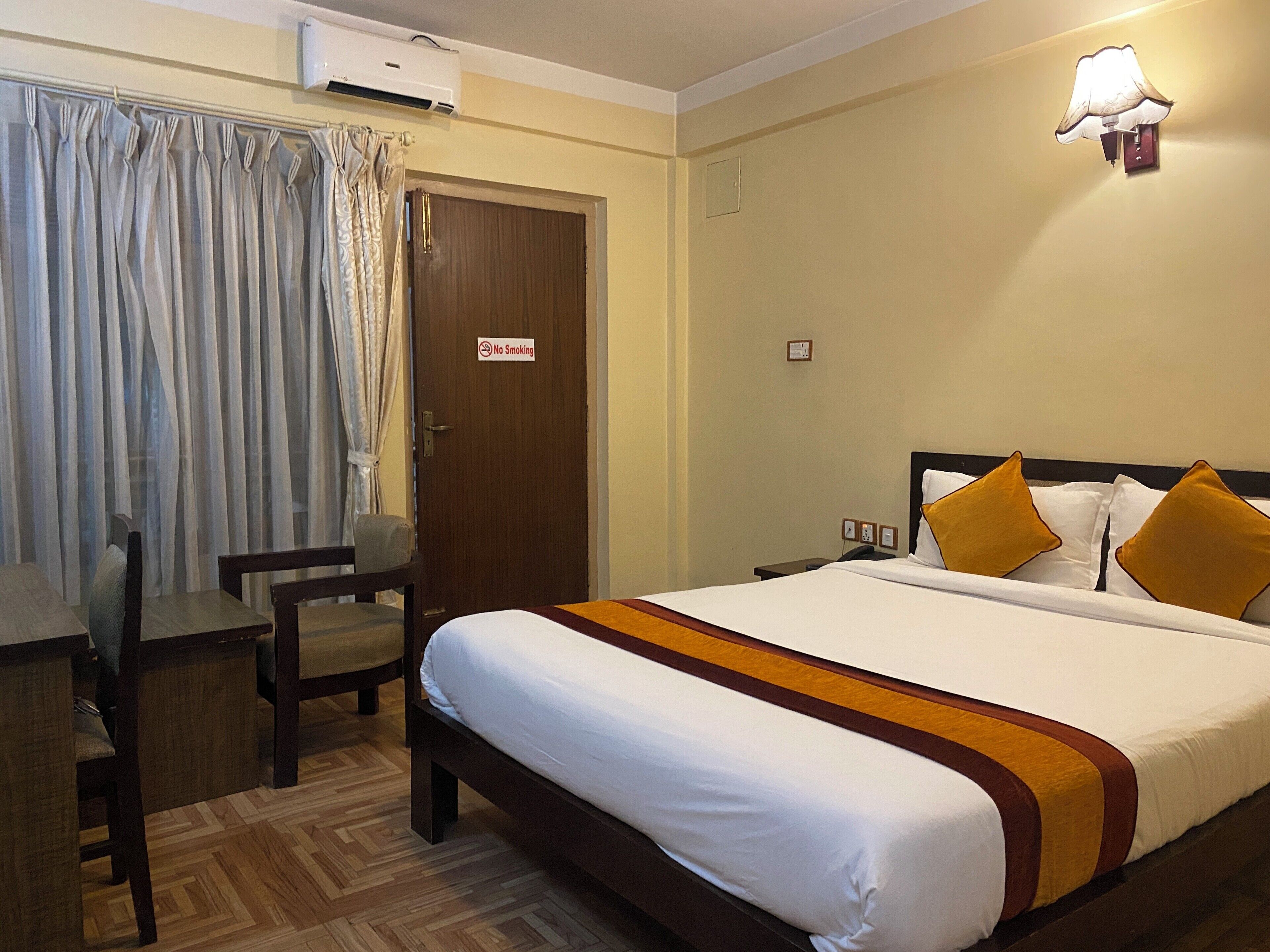 Una camera d'albergo con un letto, una scrivania e una sedia. Sul battente della porta si trova un cartello 'No Smoking'.