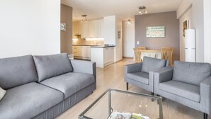 A modern living room with a grey sofa, a glass coffee table, and a dining area with a wooden table and chairs. - Beautiful Apartment With Sea-view (Blankenberge)