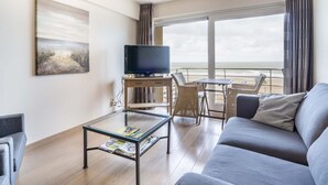 A modern living room with a sofa, a glass coffee table, a TV on a wooden stand, wicker chairs, and a large window with a view of the beach. - Beautiful Apartment With Sea-view (Blankenberge)