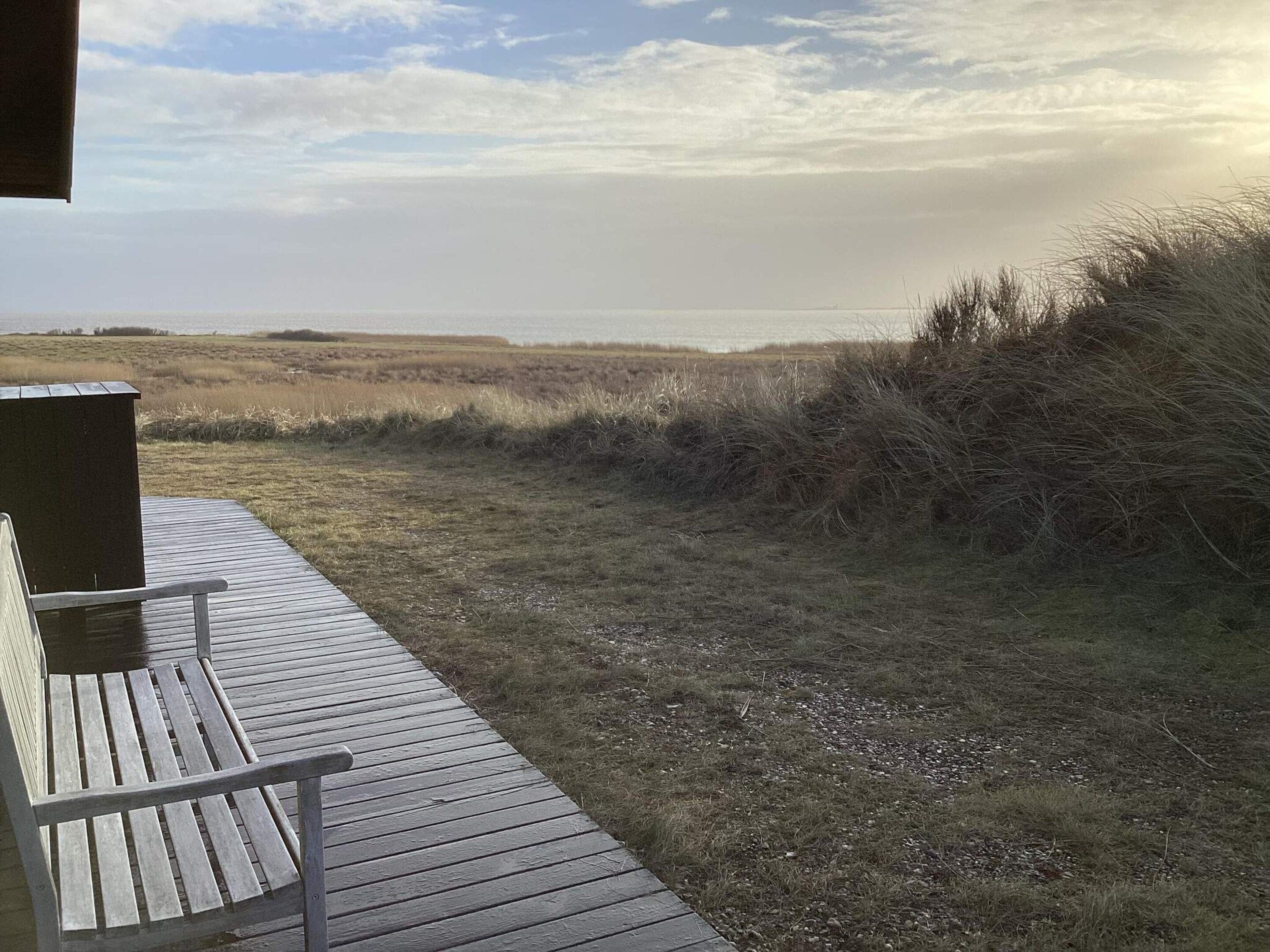 A wooden deck with a bench, overlooking a grassy area with dunes and a distant view of the sea.