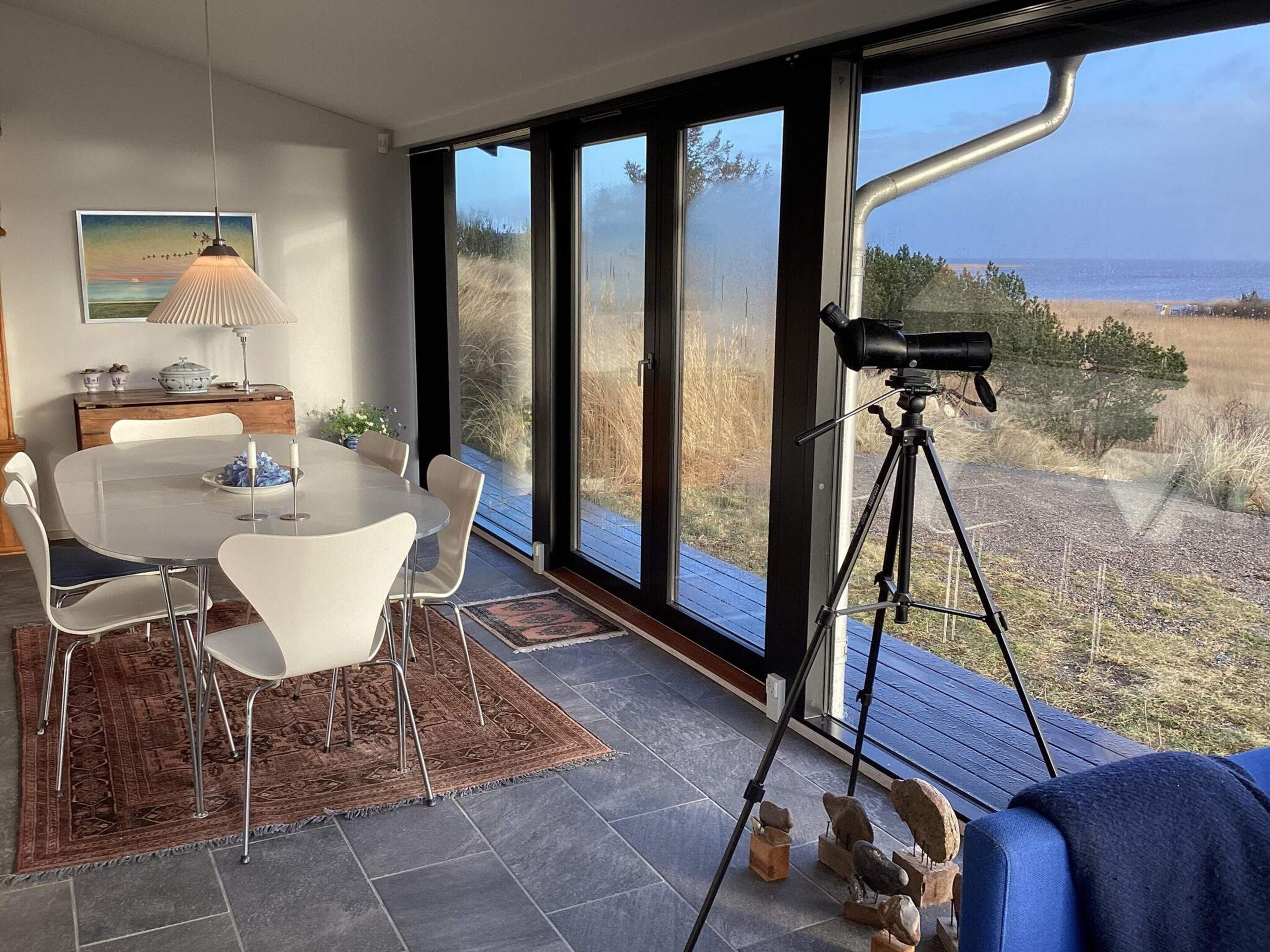 A dining area with a table and chairs, a lamp, and a view of the outdoors through large windows.