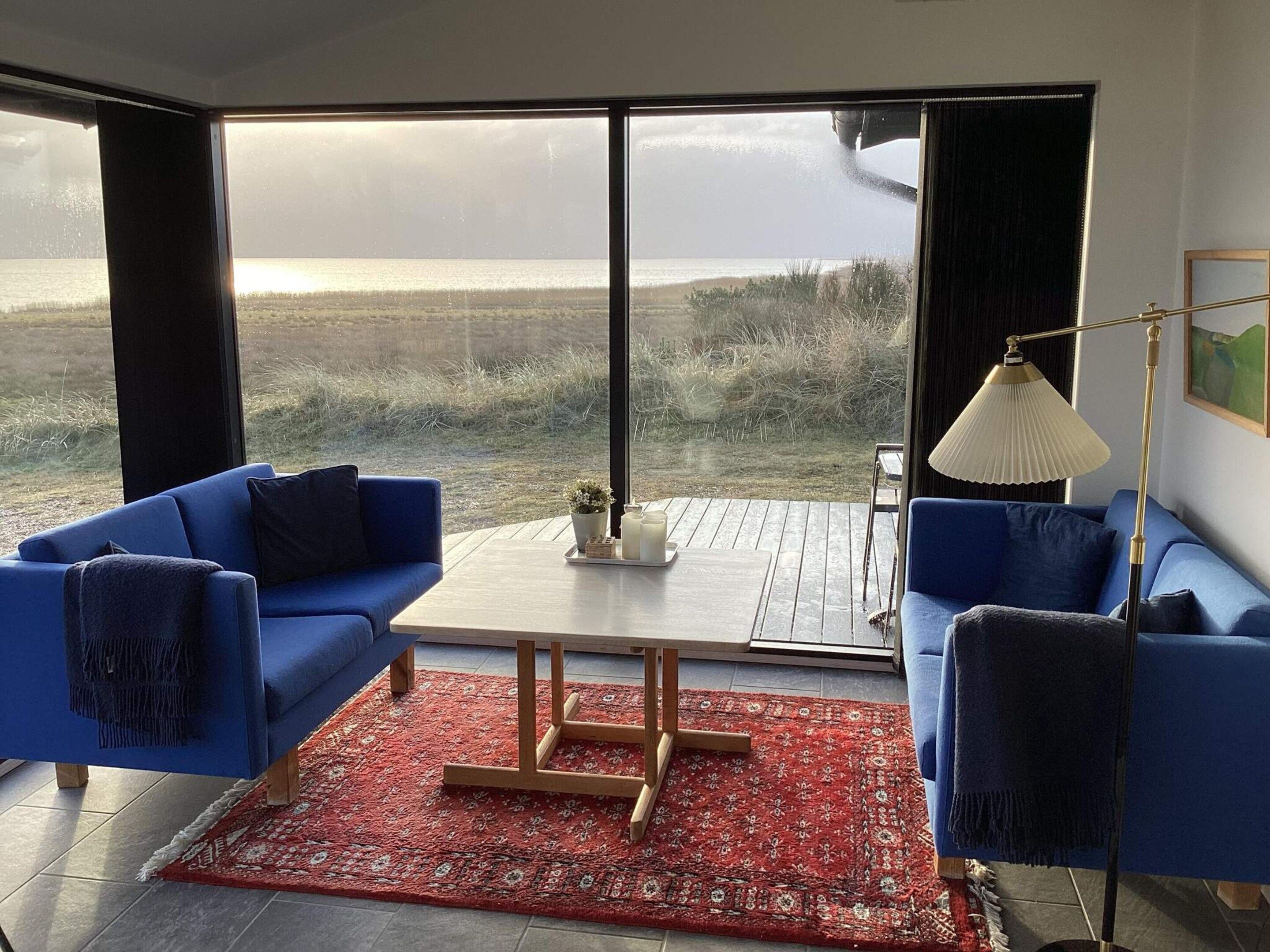 Uma sala de estar moderna com uma grande janela, dois sofás azuis, uma mesa de centro de madeira e um tapete vermelho estampado.