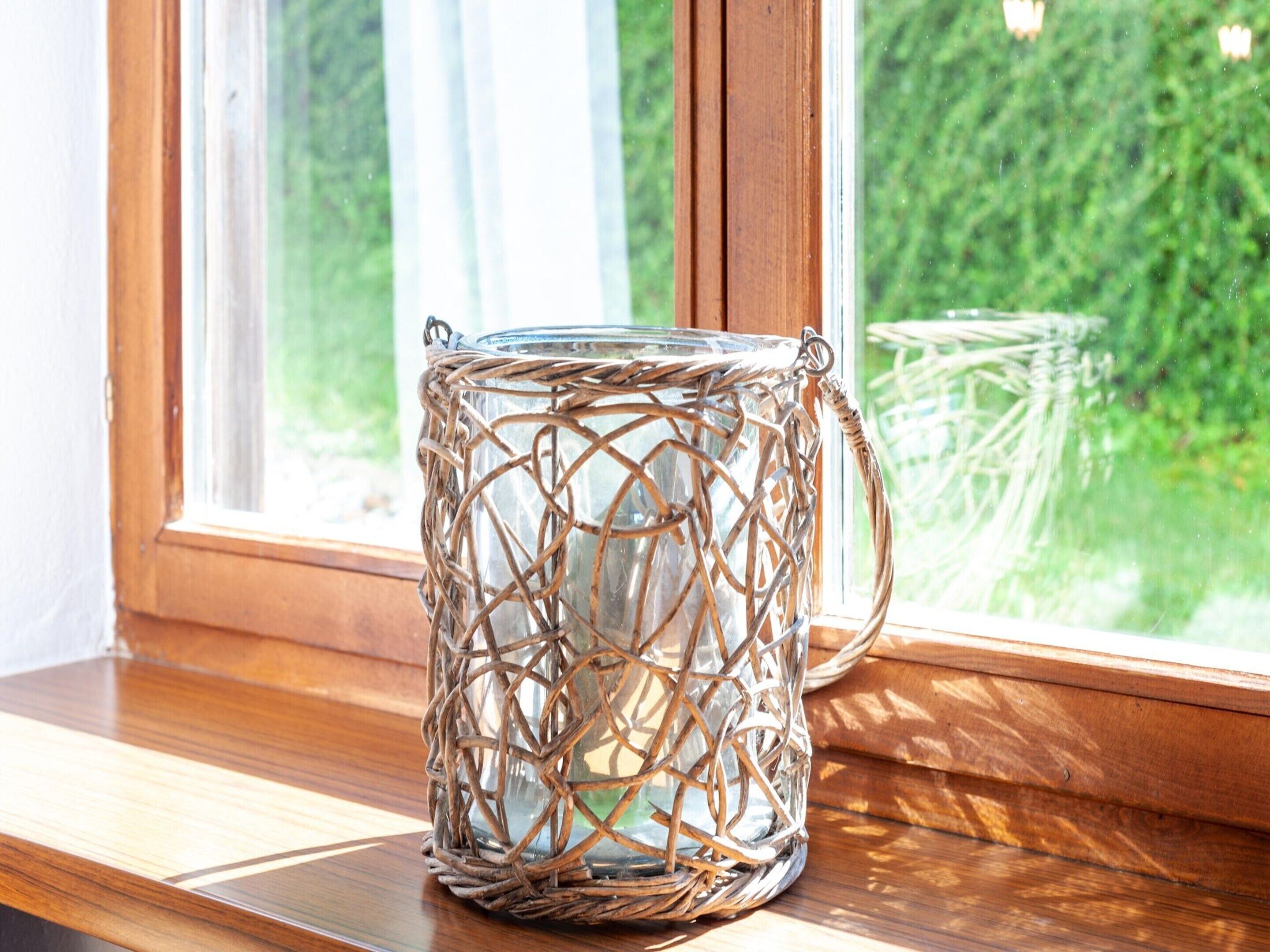 A clear glass container with a woven handle, placed on a wooden ledge by a window.