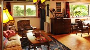 Een gezellige woonkamer met een bank, fauteuil en een tafel met een boek. Er is een raam met gordijnen, een kroonluchter en een houten plafond.